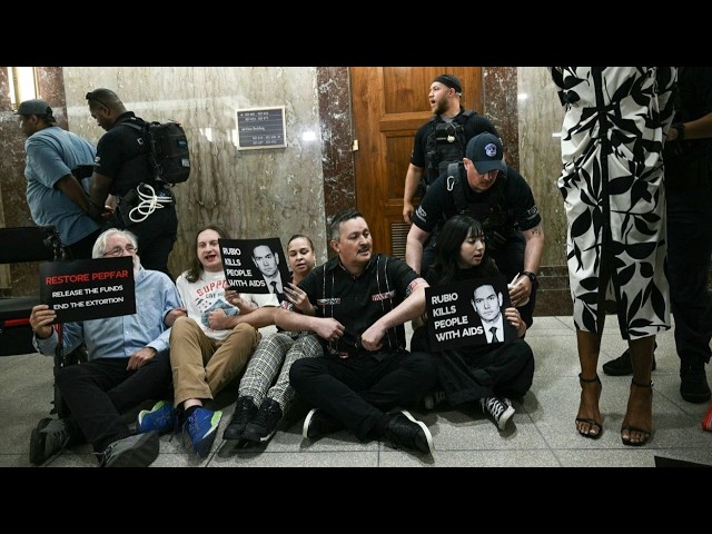 HIV/AIDS Activists Arrested After Protesting Rubio Over PEPFAR Cuts at Senate Hearing Protesters sit on the floor in a marble lobby, holding signs about funding and AIDS-related accusations as police stand nearby.