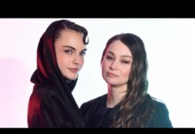 Two women pose for a studio portrait; one wears a black hooded garment while the other has long wavy hair and looks at the camera with a subtle smile.