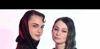 Two women pose for a studio portrait; one wears a black hooded garment while the other has long wavy hair and looks at the camera with a subtle smile.