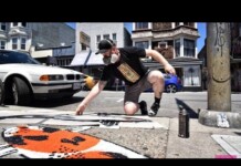 Male street artist wearing a cap and face mask crouches on a city sidewalk, spray-painting orange art with cans nearby and cars in the background