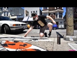 LGBTQ Artist Speaks Out After Homophobic Attacks On His Milwaukee Artwork Male street artist wearing a cap and face mask crouches on a city sidewalk, spray-painting orange art with cans nearby and cars in the background