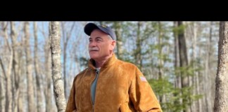 Older man with a mustache wearing a brown suede jacket and cap stands in a bare forest, looking into the distance.