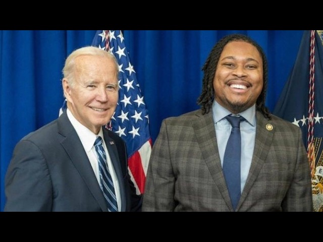 Two men in suits posing for a photo in front of American flags, smiling at the camera.
