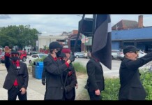 Group of men in black suits with red shirts and masks stand on a sidewalk; one salutes while a large black-and-white flag is raised nearby.