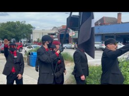 Nazi Symbols, Hate Speech Disrupt Downtown Parade Celebration at Athens Pride Group of men in black suits with red shirts and masks stand on a sidewalk; one salutes while a large black-and-white flag is raised nearby.