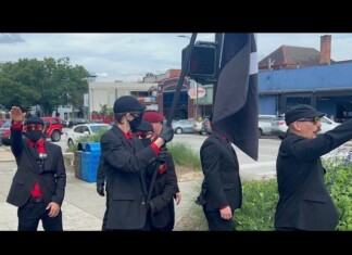 Nazi Symbols, Hate Speech Disrupt Downtown Parade Celebration at Athens Pride Group of men in black suits with red shirts and masks stand on a sidewalk; one salutes while a large black-and-white flag is raised nearby.