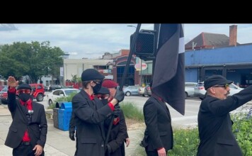Group of men in black suits with red shirts and masks stand on a sidewalk; one salutes while a large black-and-white flag is raised nearby.