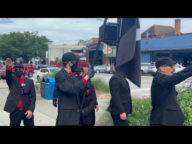 Group of men in black suits with red shirts and masks stand on a sidewalk; one salutes while a large black-and-white flag is raised nearby.