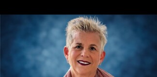 Portrait of a smiling woman with short gray hair wearing a pink scarf and necklace against a blue studio background.