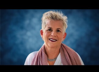 Portrait of a smiling woman with short gray hair wearing a pink scarf and necklace against a blue studio background.