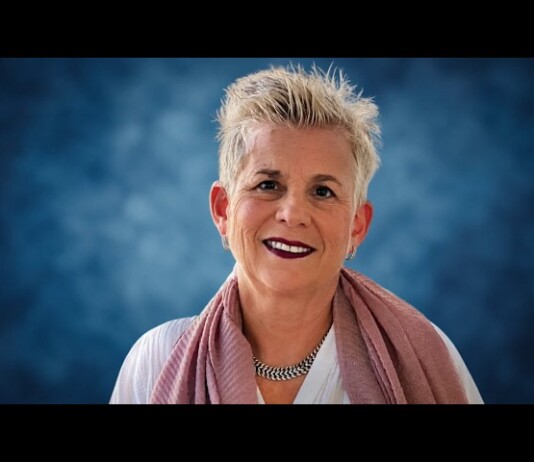 Portrait of a smiling woman with short gray hair wearing a pink scarf and necklace against a blue studio background.