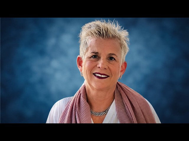 Out Former Vice Mayor Julie Carson Launches Bid for Wilton Manors Mayor Portrait of a smiling woman with short gray hair wearing a pink scarf and necklace against a blue studio background.