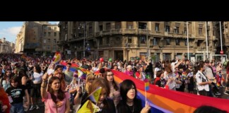 Crowd of diverse marchers at a Pride parade holding rainbow flags along a city street.