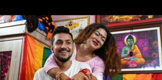 Smiling man hugged from behind by a woman in a colorful room with rainbow fabrics and framed art in the background.