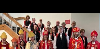 Group of clergy in red, gold, and white liturgical vestments posing for a group photo on a stairwell in a church setting.