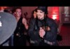 A performer with a black hat and fringed jacket claps and dances at a lively party, red ambient lighting and other revelers in the background.