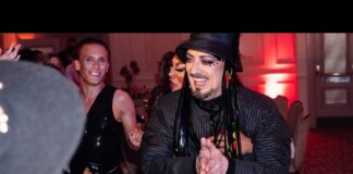 A performer with a black hat and fringed jacket claps and dances at a lively party, red ambient lighting and other revelers in the background.
