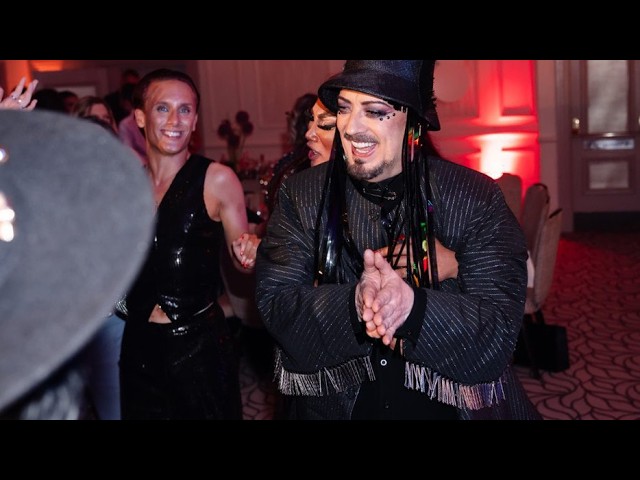 Boy George Is Honored with Lifetime Achievement Award A performer with a black hat and fringed jacket claps and dances at a lively party, red ambient lighting and other revelers in the background.