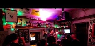 Busy bar interior at night with customers at the counter, shelves of bottles behind, and colorful neon lighting as the bartender serves drinks.