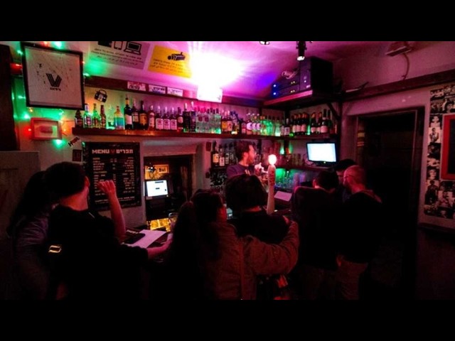 Jerusalem’s Only Gay Bar to Close After 14 Years as Owner Says Times Have Changed Busy bar interior at night with customers at the counter, shelves of bottles behind, and colorful neon lighting as the bartender serves drinks.