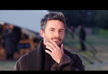 Man outdoors, thoughtful pose with hand on chin, wearing a dark brown sweater in a camp setting with tents in the background.