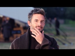 Man outdoors, thoughtful pose with hand on chin, wearing a dark brown sweater in a camp setting with tents in the background.