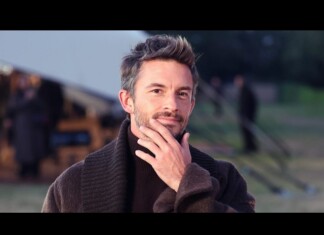 Man outdoors, thoughtful pose with hand on chin, wearing a dark brown sweater in a camp setting with tents in the background.