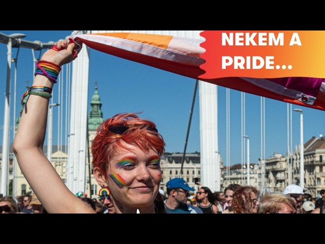 Person with rainbow makeup and colorful bracelets raises a rainbow flag at a Pride parade on a sunny city street, with a banner visible in the corner.