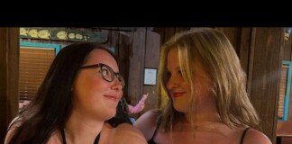 Two women sit close together at a wooden bar, smiling and looking at each other.