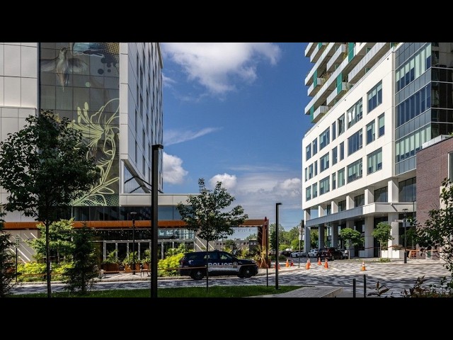 Police SUV parked at a modern urban plaza between glass office buildings, with trees and orange traffic cones nearby.