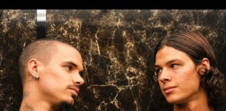 Two men face each other and share a gaze against a dark marble wall; one has a buzz cut and earrings, the other has long hair and a pendant necklace.