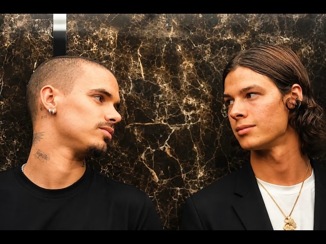 Two men face each other and share a gaze against a dark marble wall; one has a buzz cut and earrings, the other has long hair and a pendant necklace.