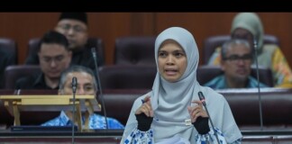 Senator Puan Marhamah Binti Rosli speaks at a parliamentary podium in a light blue hijab, with attendees in the background.