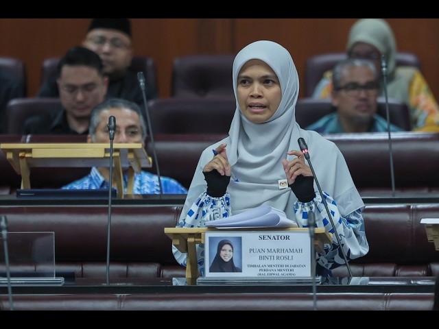 Senator Puan Marhamah Binti Rosli speaks at a parliamentary podium in a light blue hijab, with attendees in the background.