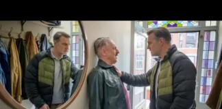 Two men in puffer jackets confront a man in a leather jacket inside a coat shop; one man rests a hand on the other's chest while they talk.
