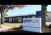 Exterior view of a modern clinic building with a blue-and-white HELP Center sign reading 'for LGBT Health and Wellness' near a tree and fall foliage.