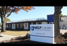 Exterior view of a modern clinic building with a blue-and-white HELP Center sign reading 'for LGBT Health and Wellness' near a tree and fall foliage.