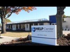 Exterior view of a modern clinic building with a blue-and-white HELP Center sign reading 'for LGBT Health and Wellness' near a tree and fall foliage.