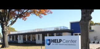 Exterior view of a modern clinic building with a blue-and-white HELP Center sign reading 'for LGBT Health and Wellness' near a tree and fall foliage.