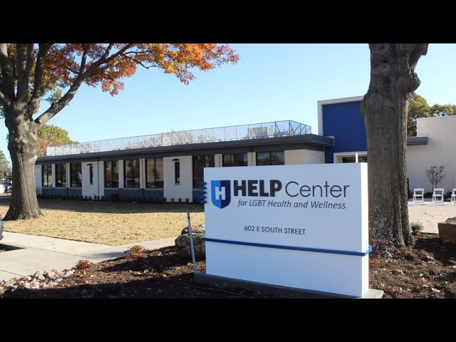 Exterior view of a modern clinic building with a blue-and-white HELP Center sign reading 'for LGBT Health and Wellness' near a tree and fall foliage.