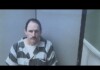 Mugshot of a man with a mustache wearing a gray and black striped prison uniform in a small interrogation room, looking at the camera.