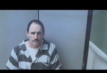 Mugshot of a man with a mustache wearing a gray and black striped prison uniform in a small interrogation room, looking at the camera.