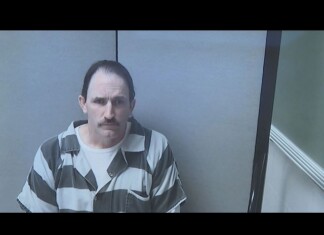 Ohio Pastor Who Opposed Pride Books Sentenced to 15 Years for Sexual Abuse of Minor Mugshot of a man with a mustache wearing a gray and black striped prison uniform in a small interrogation room, looking at the camera.