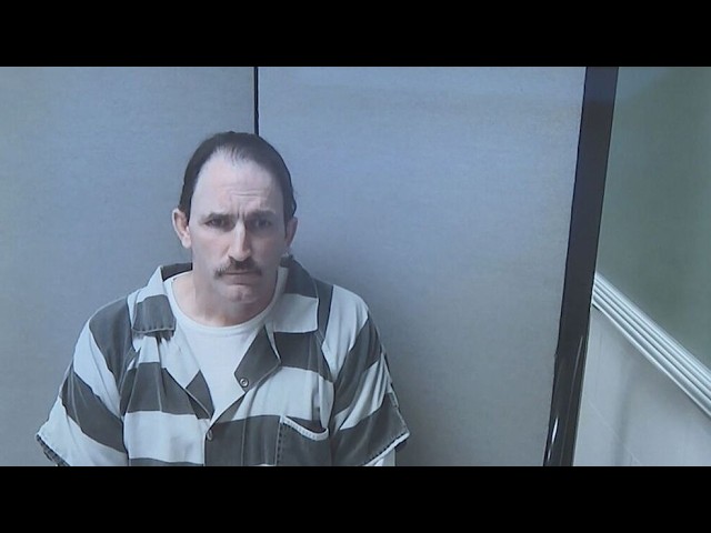 Mugshot of a man with a mustache wearing a gray and black striped prison uniform in a small interrogation room, looking at the camera.