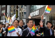 Diverse group marching in a Pride parade, waving rainbow flags along a busy city street with onlookers and storefronts behind them.