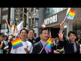 Diverse group marching in a Pride parade, waving rainbow flags along a busy city street with onlookers and storefronts behind them.