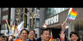 Diverse group marching in a Pride parade, waving rainbow flags along a busy city street with onlookers and storefronts behind them.