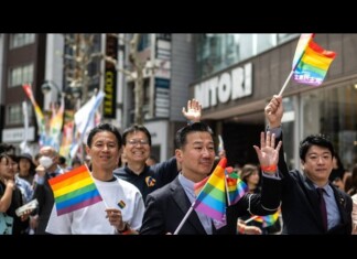 Diverse group marching in a Pride parade, waving rainbow flags along a busy city street with onlookers and storefronts behind them.
