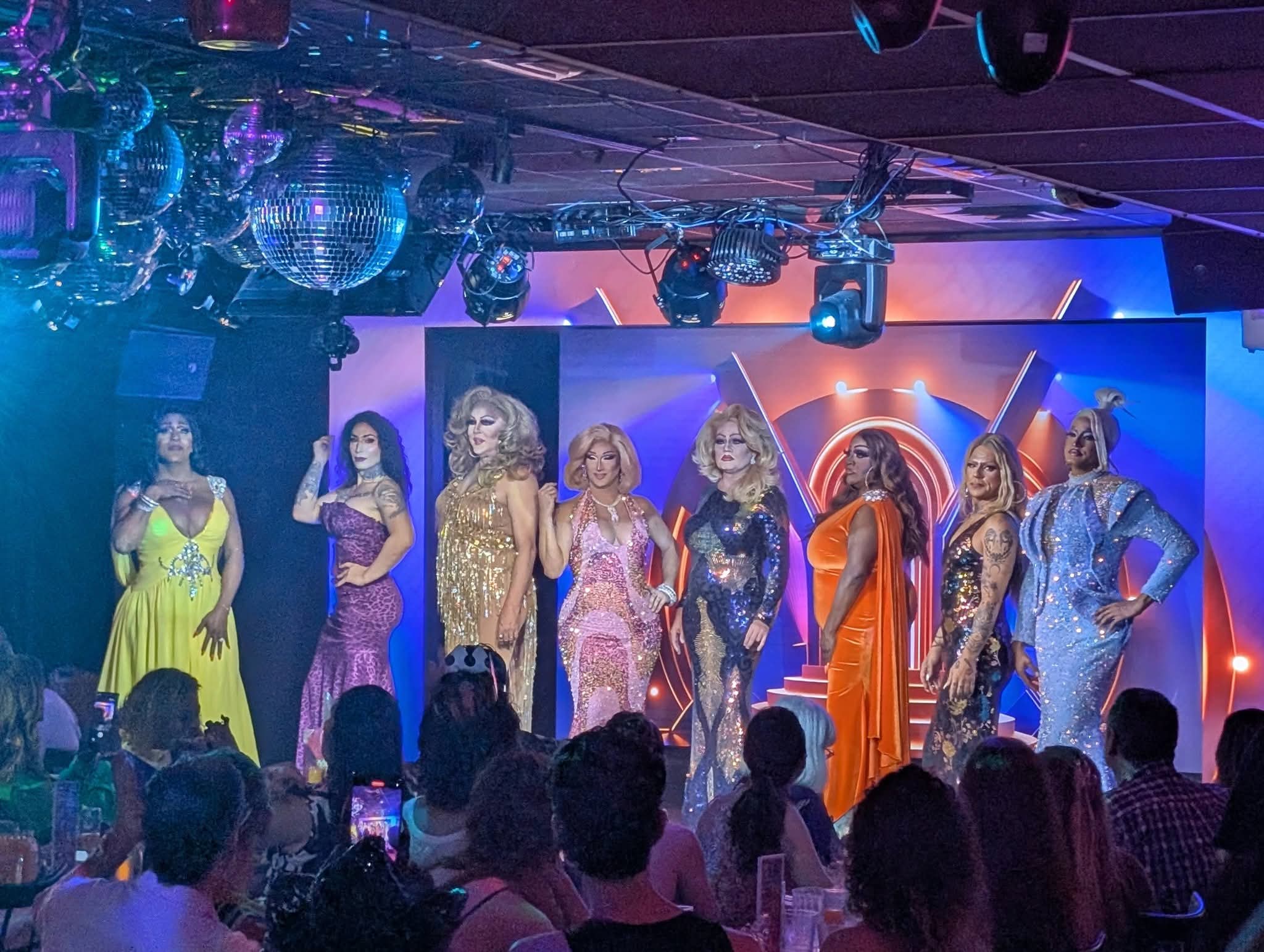 Out With Scott – By Scott Holland | Hotspots Magazine Hotspots Magazine Drag queens in glittery gowns posing on a lit stage with an audience watching.