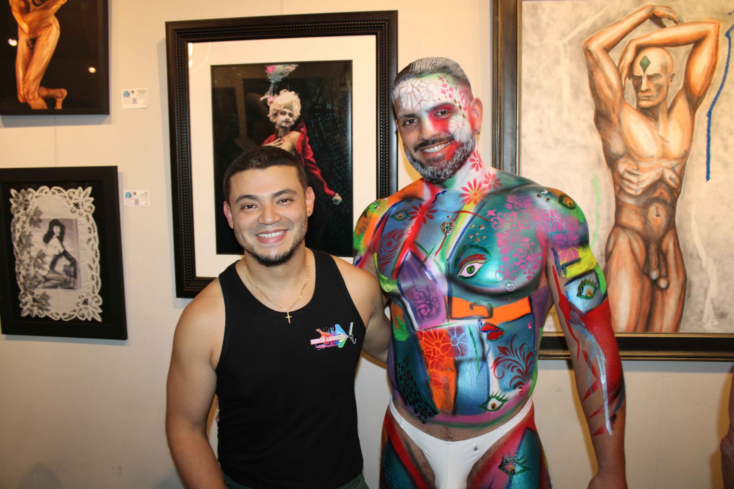 Out With Scott – By Scott Holland | Hotspots Magazine Hotspots Magazine Man in a black tank top smiling beside a second person covered in vibrant, multi‑colored body paint in an art gallery setting.